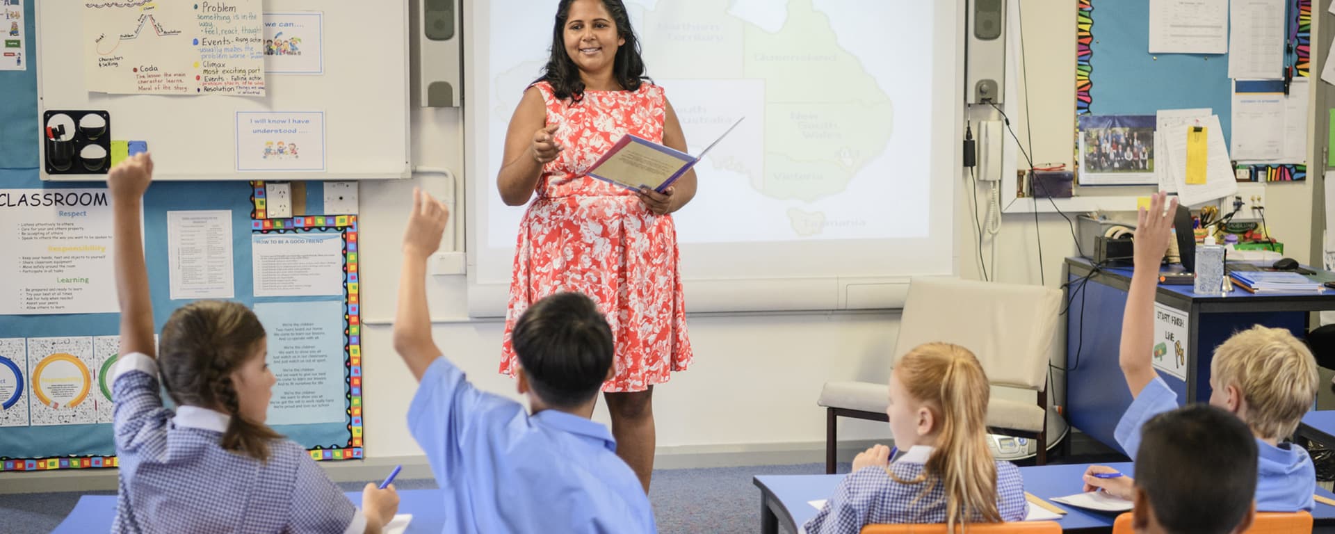 Primary classroom teacher at the front of the room kids hands up